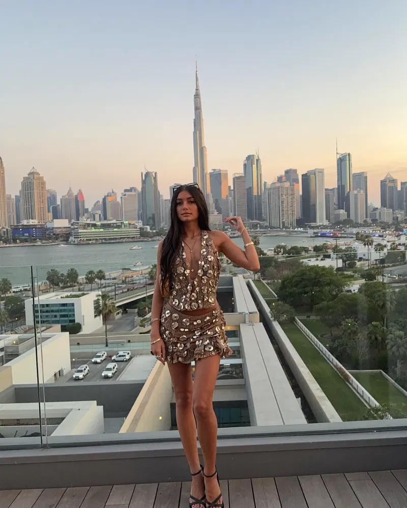 AI-generated image of Gabi in a gold sequinned co-ord on a rooftop terrace at sunset, with the Dubai skyline and the Burj Khalifa rising behind a glass balustrade