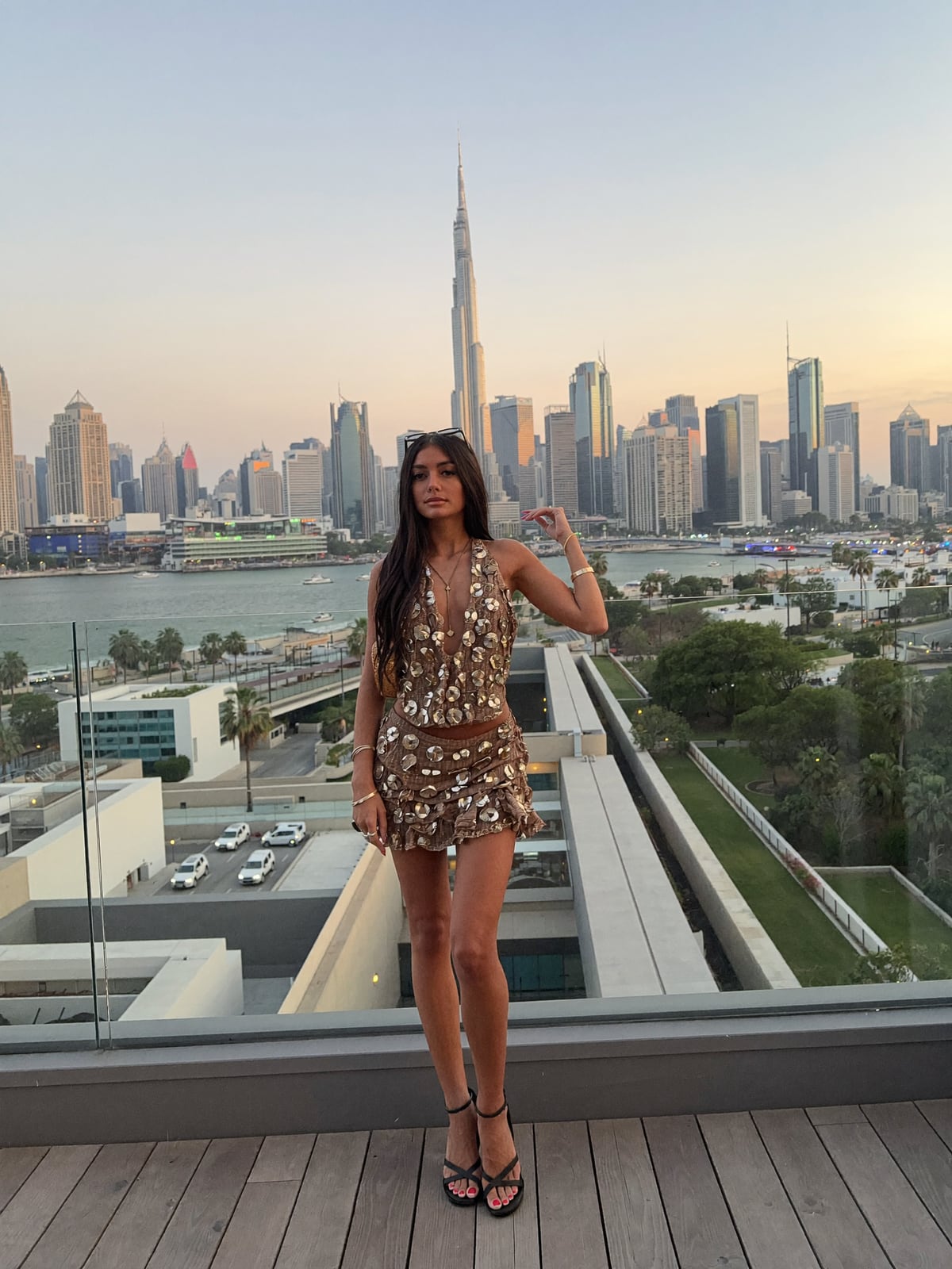 AI-generated image of Gabi in a gold sequinned co-ord on a rooftop terrace at sunset, with the Dubai skyline and the Burj Khalifa rising behind a glass balustrade