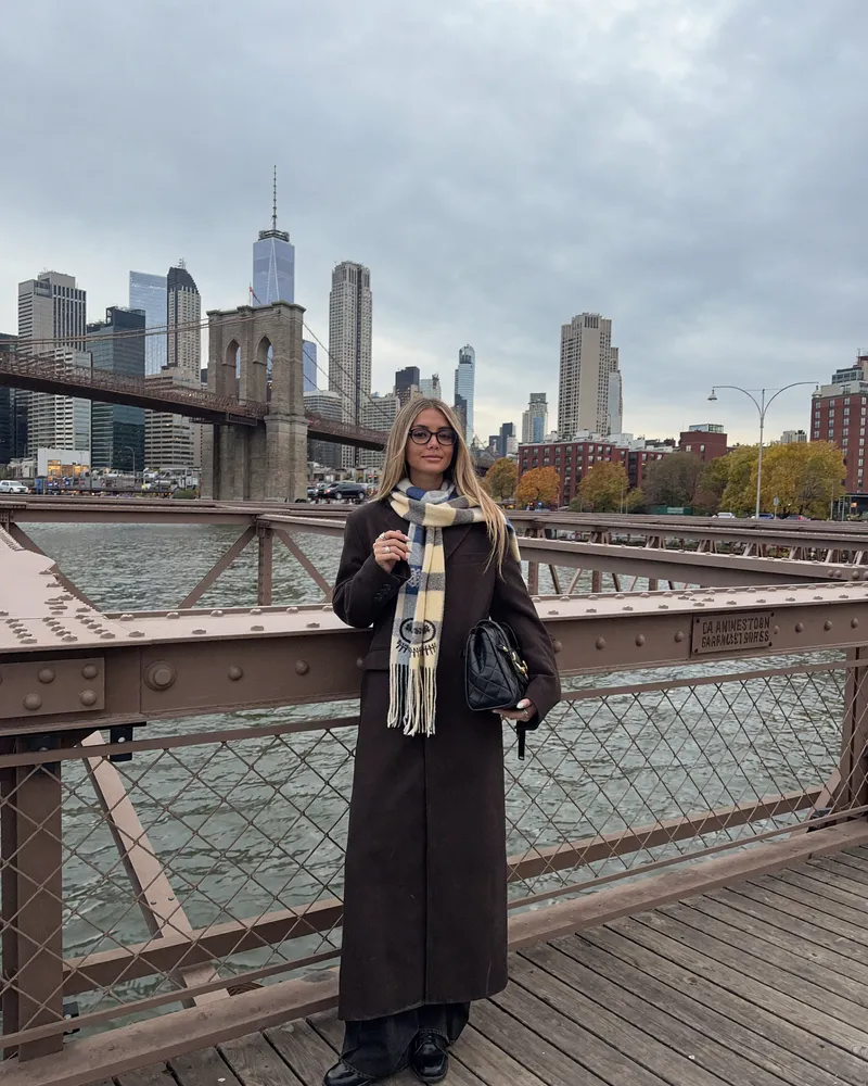 AI-generated image of Gabi in a long coat and scarf on the Brooklyn Bridge in autumn with the Manhattan skyline
