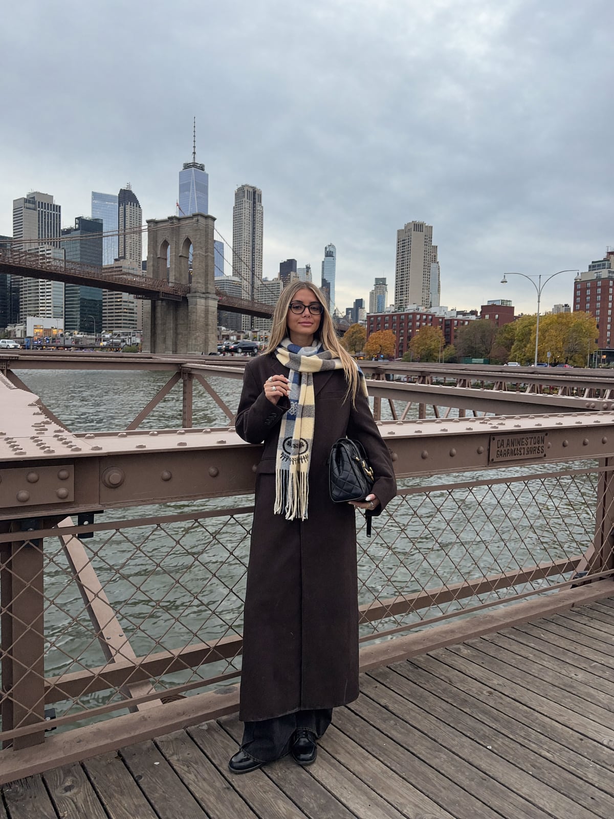 AI-generated image of Gabi in a long brown coat and plaid fringe scarf standing on the Brooklyn Bridge walkway in autumn, the Manhattan skyline and One World Trade Center behind her under an overcast sky