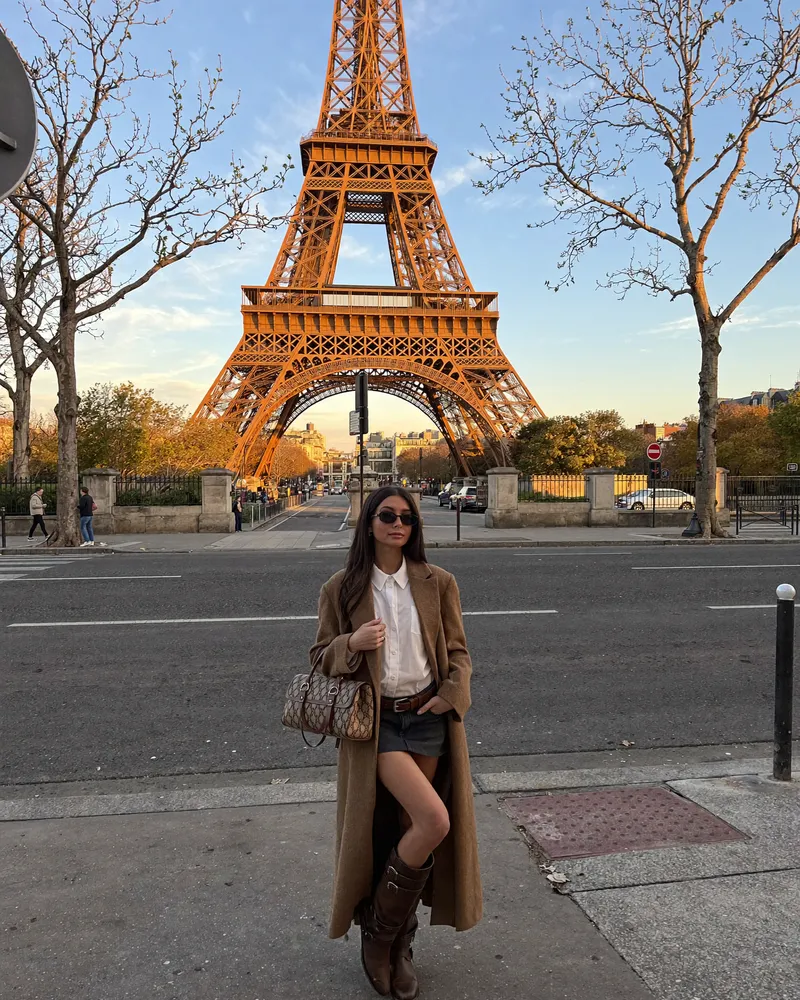 AI-generated image of Gabi in a long camel coat, white blouse and brown boots standing across the street from the Eiffel Tower lit gold at sunset, bare winter trees framing the scene