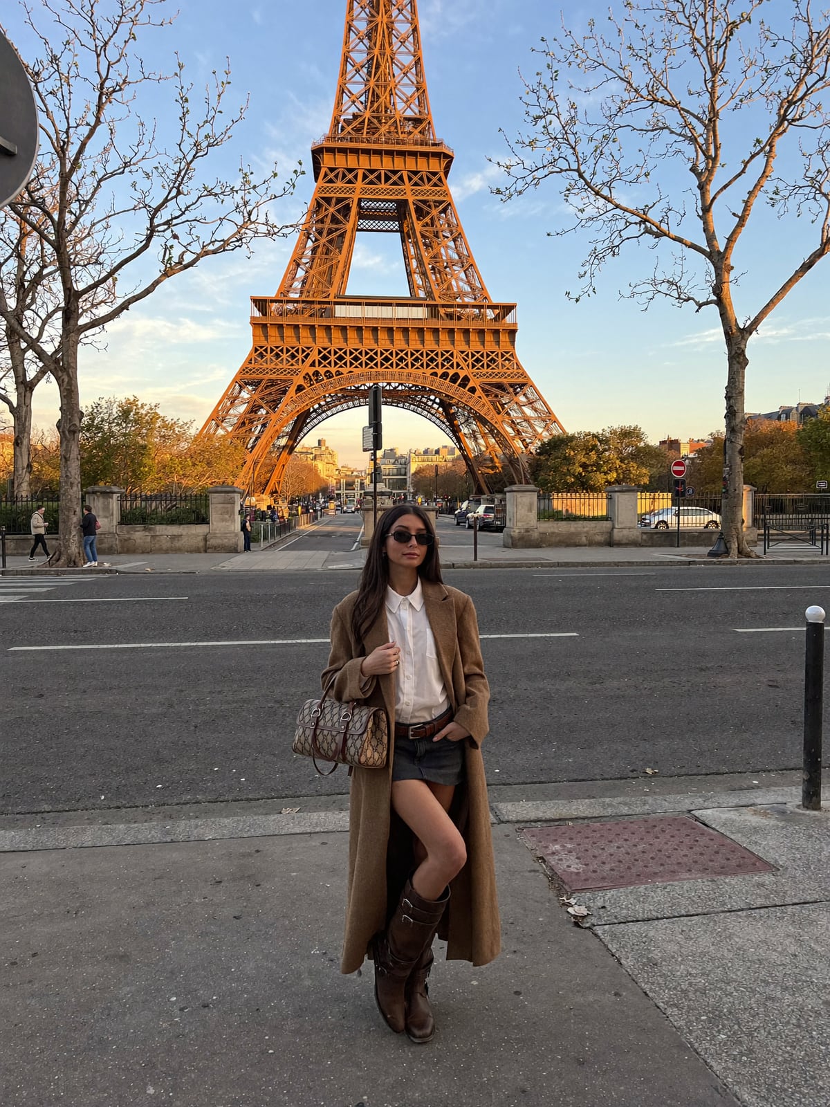AI-generated image of Gabi in a long camel coat, white blouse and brown boots standing across the street from the Eiffel Tower lit gold at sunset, bare winter trees framing the scene
