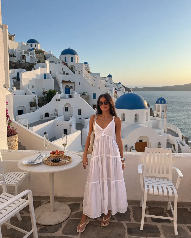 AI-generated image of Gabi in a white tiered sundress on a terrace in Oia, Santorini, with blue-domed churches and the caldera behind her at golden-hour sunset