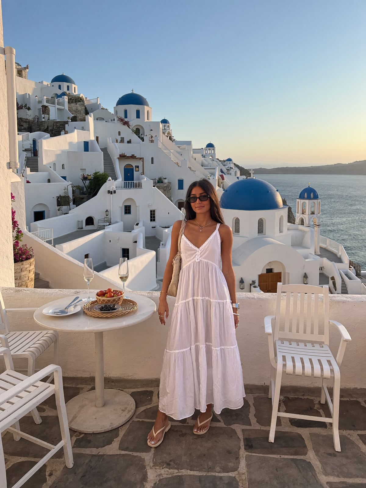 AI-generated image of Gabi in a white tiered sundress on a terrace in Oia, Santorini, with blue-domed churches and the caldera behind her at golden-hour sunset