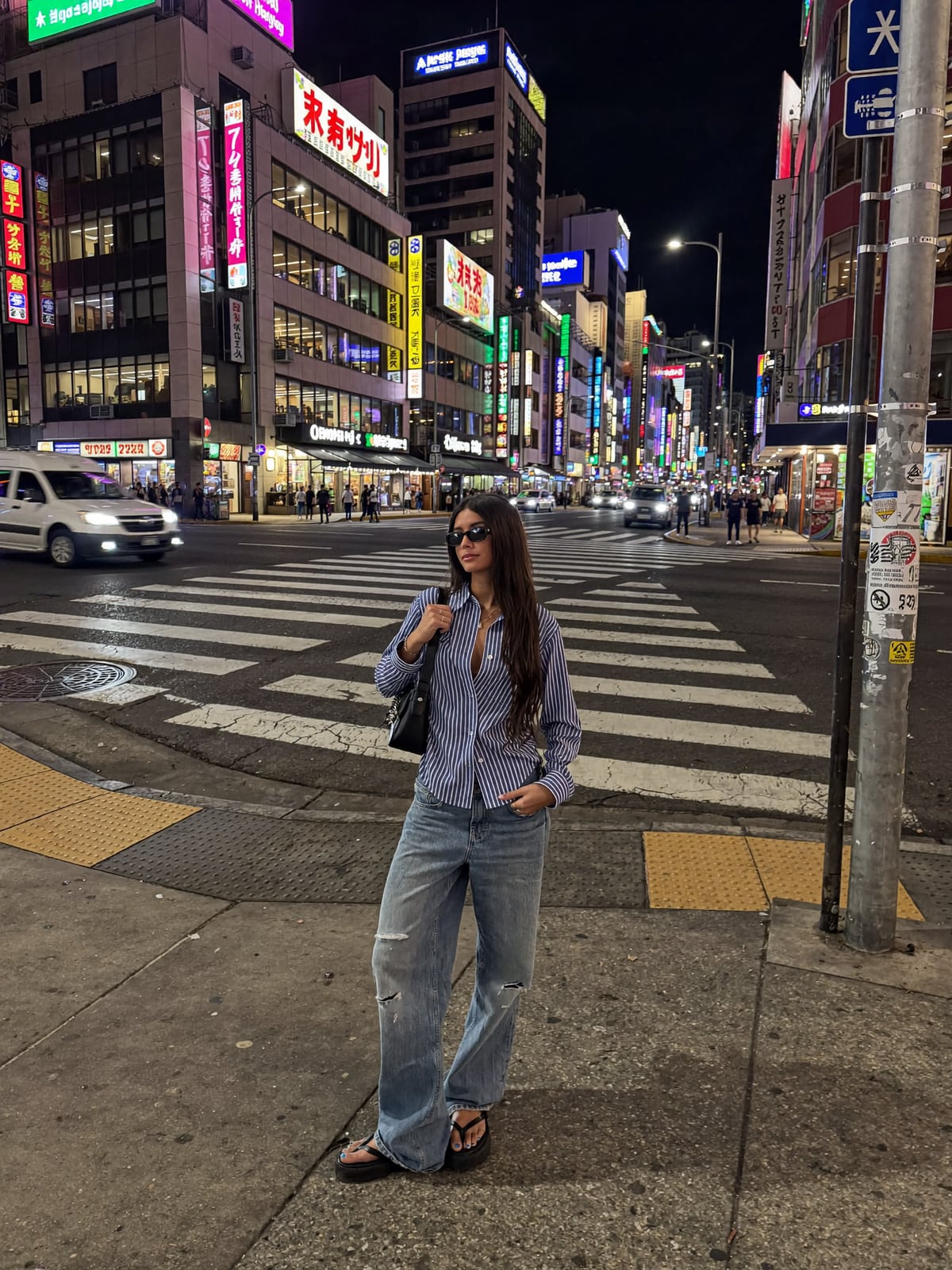 AI-generated image of Gabi in a striped shirt and light-wash wide-leg jeans standing at a neon-lit Shinjuku street crossing in Tokyo at night, towering Japanese signage glowing behind her